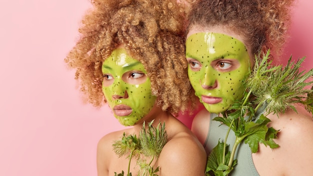 Horizontal shot of curly haired young female models apply green\
facial masks for skin treatment use natural organic products have\
thoughtful expressions isolated over pink background copy\
space
