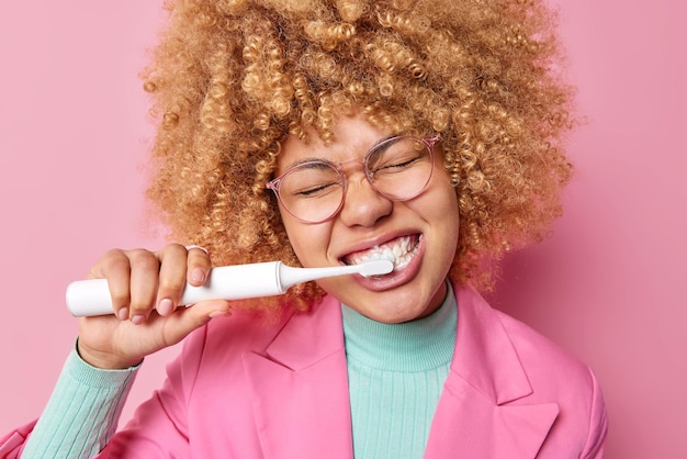 Горизонтальный снимок кудрявой молодой женщины, чистящей зубы каждое утро, пытается быть всегда здоровой, использует электрическую зубную щетку, носит прозрачные очки, формальную одежду, изолированную на розовой стене.