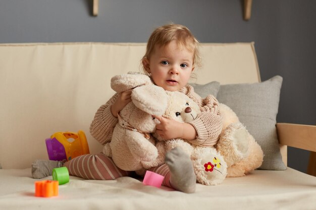 Horizontal shot of cute blonde baby girl in beige jumper sitting on sofa with a plush big toys looking at camera playing alone at home posing among multicolored sorter parts