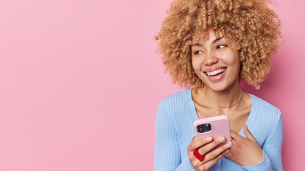 Horizontal shot of cute curly haired woman holds mobile phone checks received email message browses website in social media wears casual blue jumper isolated over pink background copy space