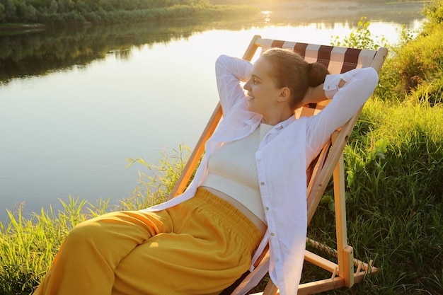 Horizontal shot of cute dark haired Caucasian woman wearing white shirt and yellow trousers sitting in the folding chair by the water raised arms looking at beautiful views