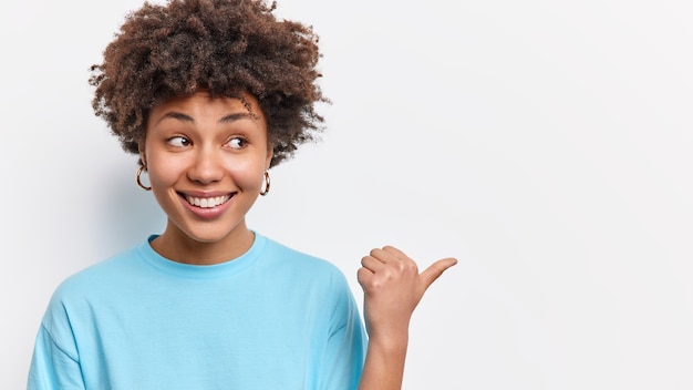 Horizontal shot of cute pleased Afican American woman shows store promo deal smiles positively points at right copy space isolated over white wall shares good news with you. Check this out