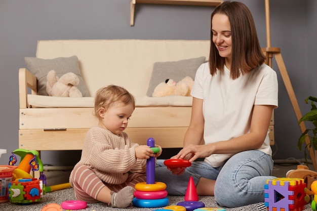 Horizontal shot of dark haired woman sitting on floor and playing with her baby daughter with pyramid kid learning colors and sizes early development