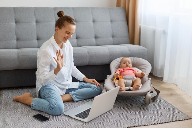 Horizontal shot of delighted woman with bun hairstyle wearing white shirt and jeans sitting on floor with hey newborn baby and having video call on laptop waving hand saying hello