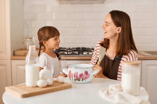 Horizontal shot of delighted young adult woman cooking with her daughter in the kitchen, having fun together, laughing, looking at each other with happy facial expression.