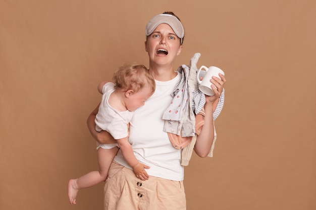 Horizontal shot of desperate sad frustrated mother with baby daughter and laundry in hands being tired needs rest crying expressing sorrow posing isolated over brown background