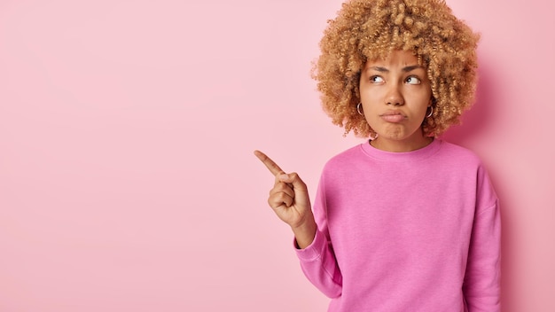 Horizontal shot of displeased curly haired woman shows\
something uninteresting points index finger at blank space doesnt\
like something dressed in casual clothes isolated over pink\
background