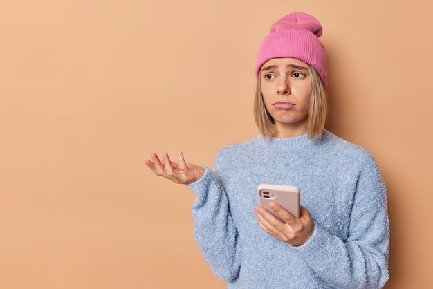 Horizontal shot of displeased puzzled young woman looks sadly away shrugs shoulders holds mobile phone cannot receieve necessary call wears casual jumper and hat isolated over beige background