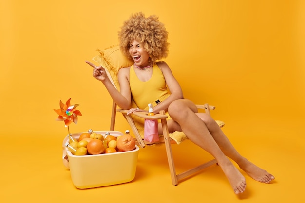 Horizontal shot of emotional woman with curly hair exclaims\
loudly points index finger away notices something sunbathes at deck\
chair dressed in swimwear uses portable fridge isolated on yellow\
wall