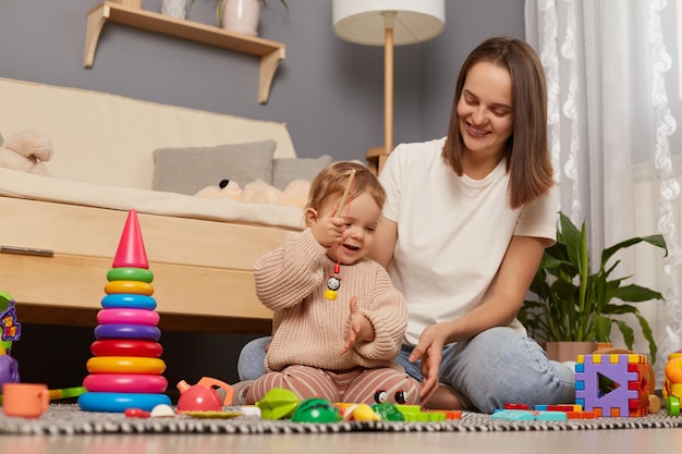 Horizontal shot of excited cheerful joyful kid playing with magnet toys sitting with her mother on floor expressing happiness play developing games spending time at home