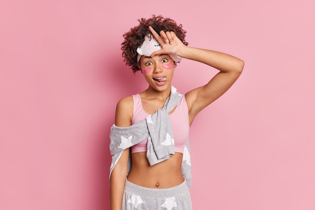 Photo horizontal shot of funny woman makes loser gesture sticks out tongue teases someone dressed in sleepwear wears beauty pads isolated over pink wall