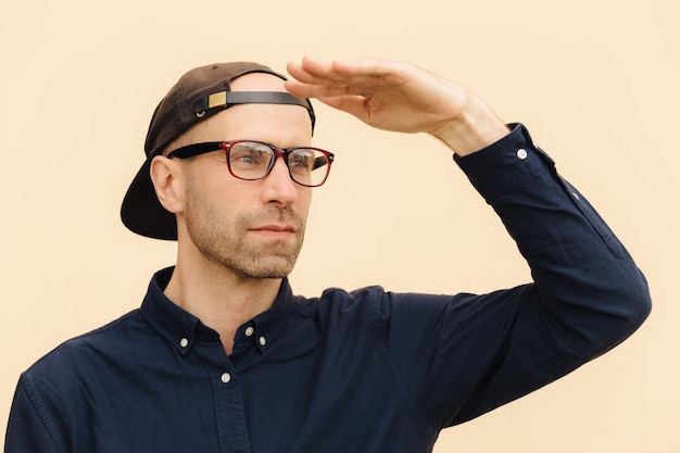 Horizontal shot of handsome male looks thoughtfully into distance keeps hand near forehead wears spectacles cap and shirt poses against light studio background Pensive dreamy man indoor