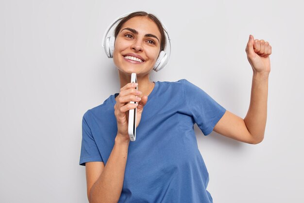 Horizontal shot of happy brunette woman dances carefree keeps smartphone near mouth as if microphone uses stereo headphones enjoys favorite music wears blue t shirt isolated over white background