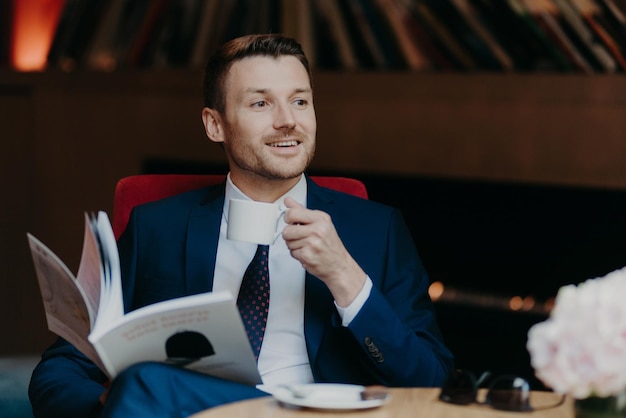 Horizontal shot of happy businessman reads popular magazine in
cafeteria drinks aromatic coffee dressed in formal suit has
pleasant smile wears formal black suit enjoys recreation time