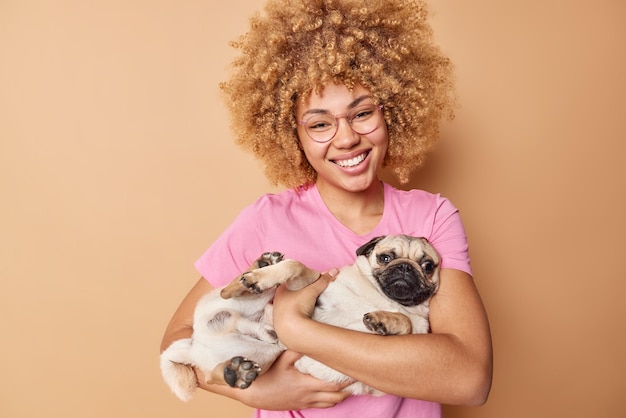 Inquadratura orizzontale di felice donna dai capelli ricci posa con adorabile cane carlino sdraiato sulle mani sorride positivamente vestito casualmente isolato su sfondo beige il cucciolo trascorre del tempo con il suo amorevole proprietario