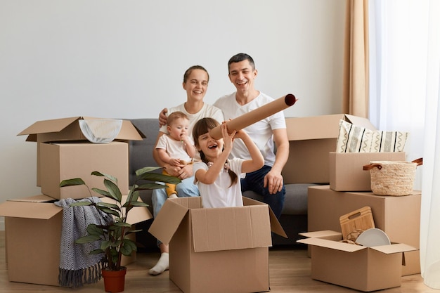 Horizontal shot of happy family relocating in new flat and having fun together while unpacking their personal belongings little daughter sitting in cardboard box with handmade spyglass