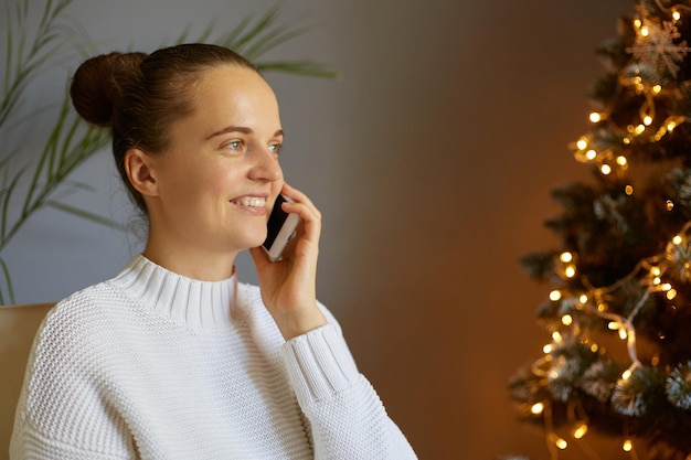 Horizontal shot of happy joyful attractive woman with bun hairstyle wearing white sweater standing at home near Christmas tree talking on mobile phone smiling happily