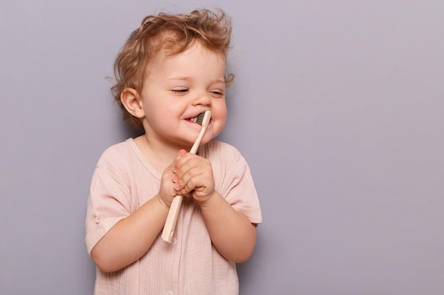 Горизонтальный снимок счастливой девочки, чистящей зубы, держащей зубную щетку в руках, пытающейся сделать это самостоятельно, стоя изолированной на сером фоне, копируя пространство для рекламы