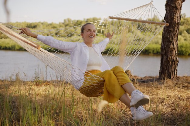 Horizontal shot of happy laughing woman sitting on hammock by the water expressing happiness enjoying weekend in beautiful nature with excellent view funny rest