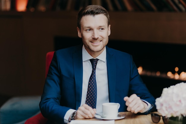 Horizontal shot of happy male office worker has coffee break at cafeteria drinks hot beverage dressed in formal clothing sits in restaurant looks joyfully into distance focused somewhere