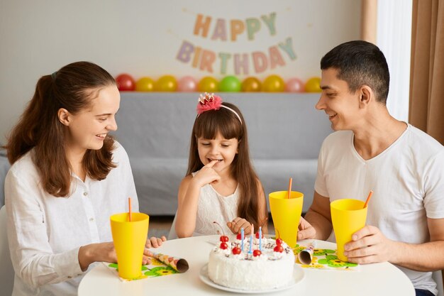 Horizontal shot of happy optimistic woman man and child sitting at table and eating birthday cake expressing positive emotions being in good mood balloons and party inscription on background