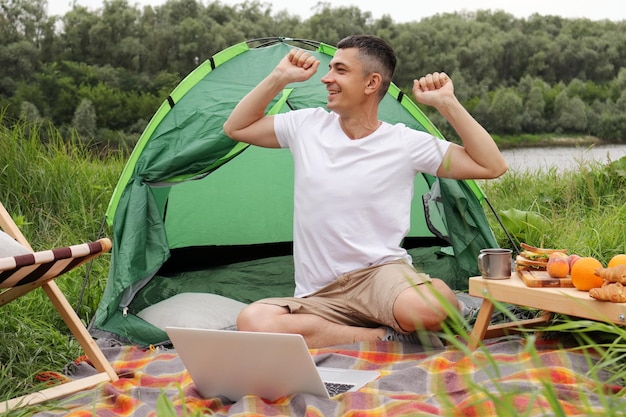 Horizontal shot of happy positive man freelancer spending time in nature on camping waking up early for working online enjoying time in open air stretching hands looking away