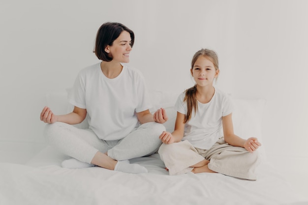 Horizontal shot of happy relaxed brunette mother and daughter pose in lotus pose try to relax keep legs crossed dressed in casual white clothes meditate on bed pose against light background