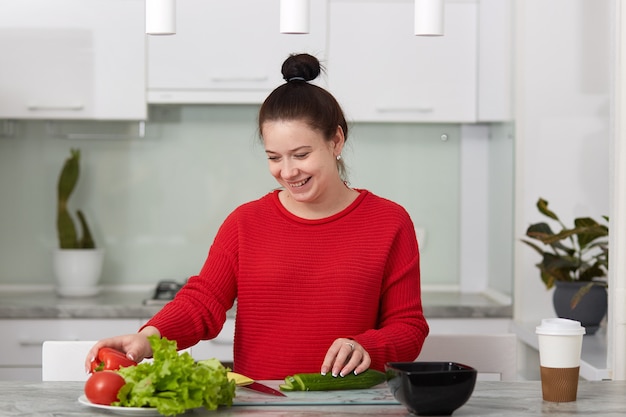 Horizontal shot of happy smiling pregnant woman cuts vegetables for making salad, poses against kitchen interior, wears red casual sweater, cute female waits for child. Motherhood and children concept