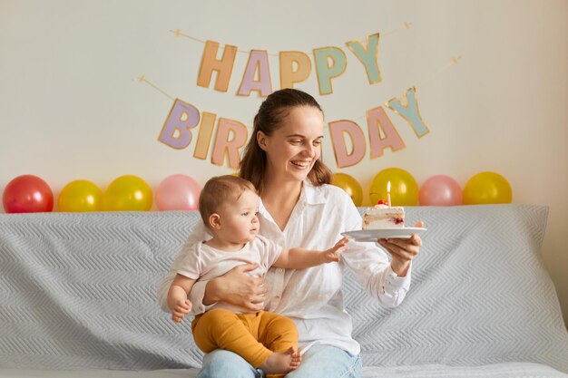 Horizontal shot of happy smiling woman sitting on sofa with her infant daughter and holding cake with candles, smiling happily, expressing positive emotions, celebrating first baby birthday.