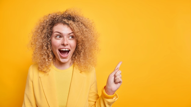 Horizontal shot of happy woman with curly bushy hair points away on blank space recommends 