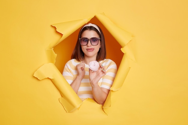 Horizontal shot of hungry positive woman wearing hair band and striped t shirt holding sugary marshmallow and showing tongue out standing in yellow paper hole