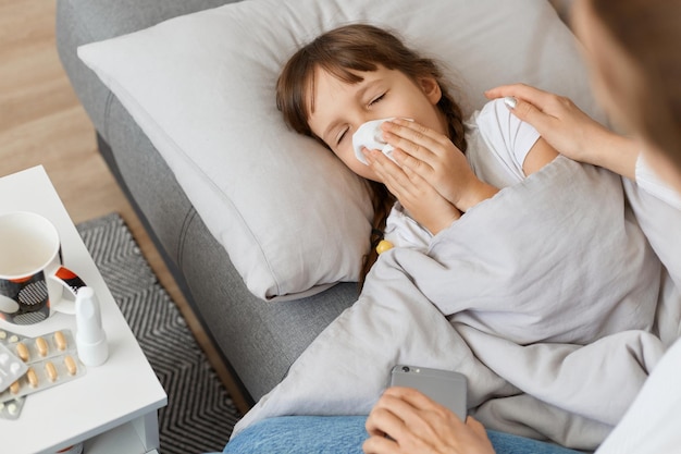 Foto inquadratura orizzontale di una bambina malata sdraiata sul divano sotto la coperta e che starnutisce madre senza volto seduta vicino ai sintomi dell'influenza del virus della figlia malata