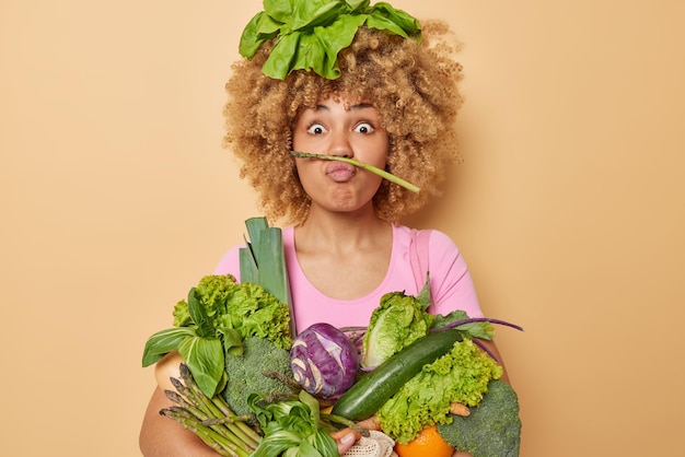 Horizontal shot of impressed curly haired young woman keeps asparagus on rounded lips carries fresh vegetables foolishes around eats organic healthy food isolated over brown studio background