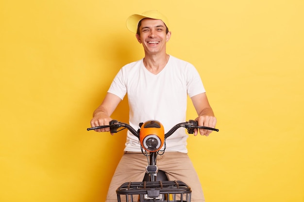 Horizontal shot of joyful Caucasian man wearing white t shirt and cap having fun riding scooter spending his free time with in active way isolated on yellow background