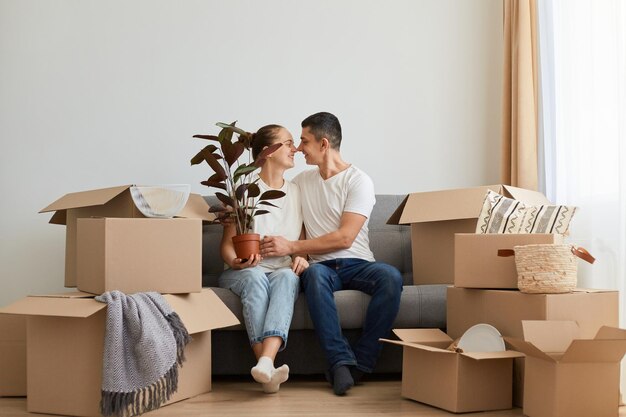 Horizontal shot of lovely romantic young family buying new flat and posing surrounded with boxes with personal pile sitting on sofa and kissing celebrating relocation into a new flat