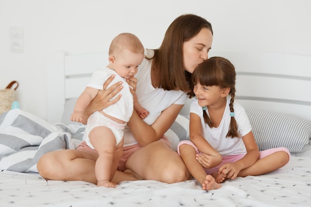 Horizontal shot of lovely woman wearing white shirt and short sitting on bed with her daughters mother holding infant baby and kissing her elder child