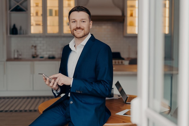 Horizontal shot of man employee uses great business app reads good news message holds mobile phone poses against kitchen interior near desktop wears formal outfit smiles happily Mobile payments