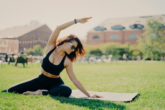 Horizontal shot of motivated young female model has regular morning workout raises arm and leans at different sides dressed in sportswear poses on karemat outside stays in good physical shape