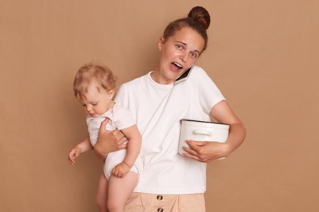Фото Горизонтальный снимок взволнованной шокированной женщины в белой рубашке в стиле кэжуал, держащей на руках младшую дочь и горшок во время разговора по смартфону на коричневом фоне