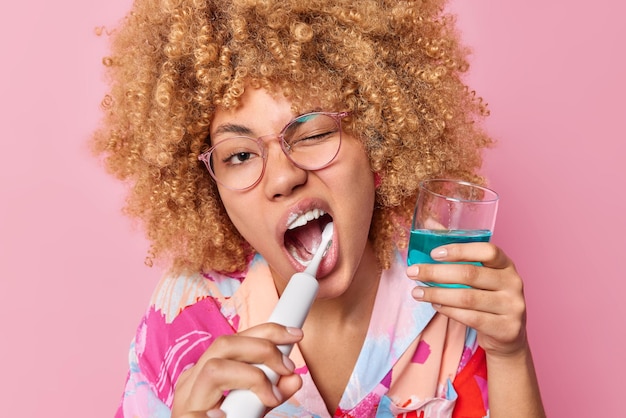 사진 곱슬머리 젊은 여성이 양치질을 하는 수평 샷은 정기적으로 구강청결제 한 잔을 들고 입을 크게 벌리고 분홍색 배경에 격리된 투명한 안경을 착용합니다. 구강 위생