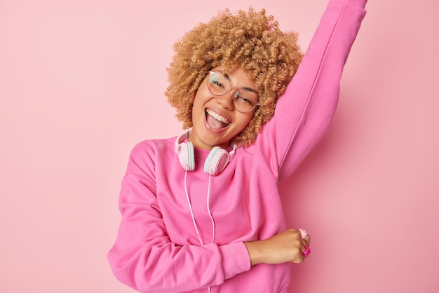 Horizontal shot of optimistic woman dances carefree foolishes\
around has fun listens favorite music wears pullover headphones\
around neck keeps arm raised smiles cheerfully isolated over rosy\
wall