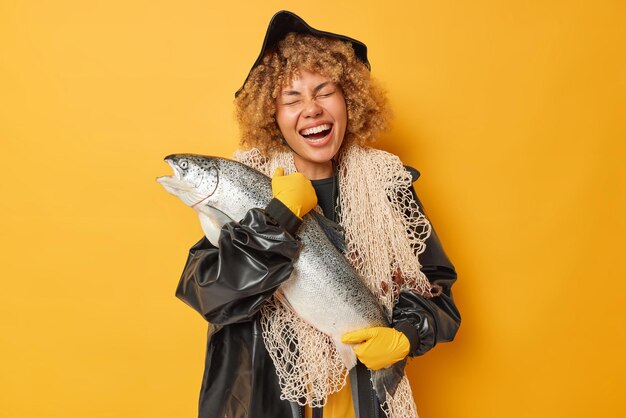 Horizontal shot of overjoyed curly haired female angler carries\
big trophy caught salmon laughs happily wears black leather\
raincoat and hat carries fishing net isolated over yellow\
background