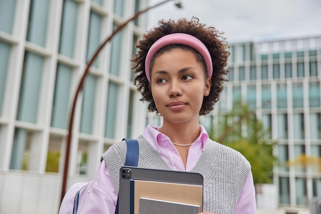 Horizontal shot of pensive millennial girl has curly hair holds notepads digital tablet carires backpack wears casual clothes poses against blurred city duing daytime