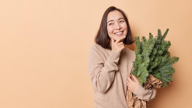 Horizontal shot of petty Asian woma holds chin looks with tender expression away holds bouquet of green spruce branches prepares for New Year isolated over beige background blank space on left