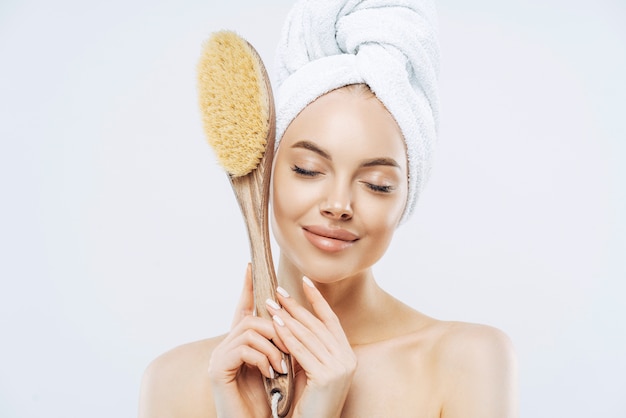 Horizontal shot of pleased female stands with eyes closed, holds big bath brush for massaging skin, enjoys hygienic treatments at home, isolated on white background. Beauty, care, exfoliation