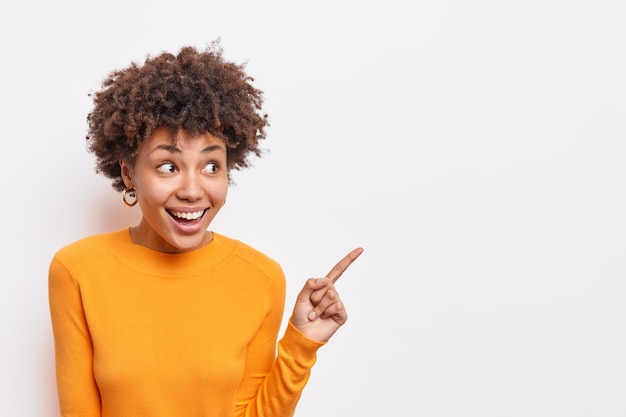 Horizontal shot of positive dark skinned woman looks with interest and amazement aside indicates at copy space shows good deal recommends product wears orange jumper isolated on white  wall