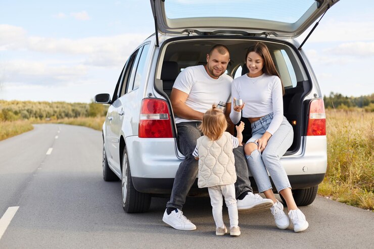Photo horizontal shot of positive delighted family with infant kid drinking hot tea from thermos sitting in car trunk stop their automobile for having rest