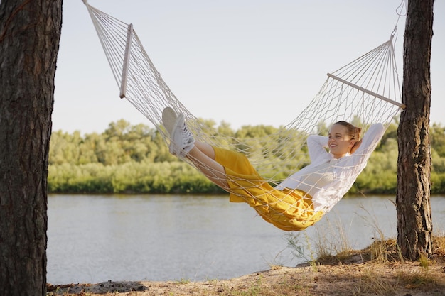 Horizontal shot of positive happy optimistic woman wearing\
white shirt sitting in hammock on the bank of the river lying with\
relaxed expression looking away with charming smile