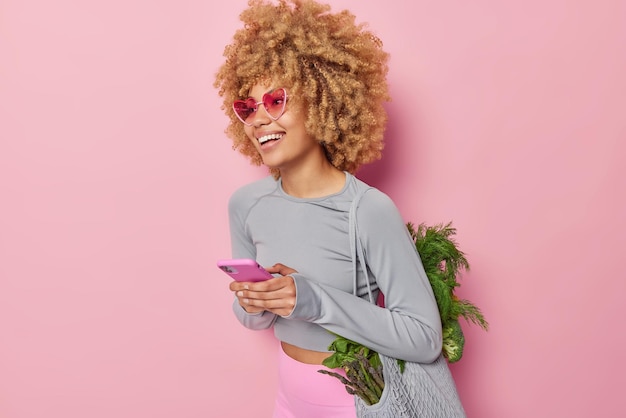 Horizontal shot of positive woman uses mobile phone for surfing internet carries bag full of fresh green vegetables returns from market wears sunglasses and activewear isolated over pink wall