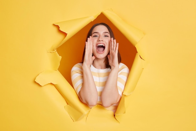 Horizontal shot of positive woman wearing striped t shirt posing standing through yellow paper torn hole screaming announcing advertisement with optimistic expression